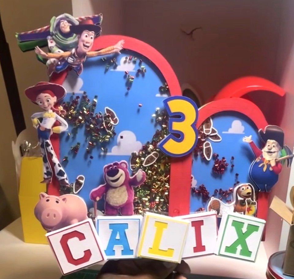 A layered shaker centerpiece with cut character figures, confetti-filled windows and blocks spelling the name Calix