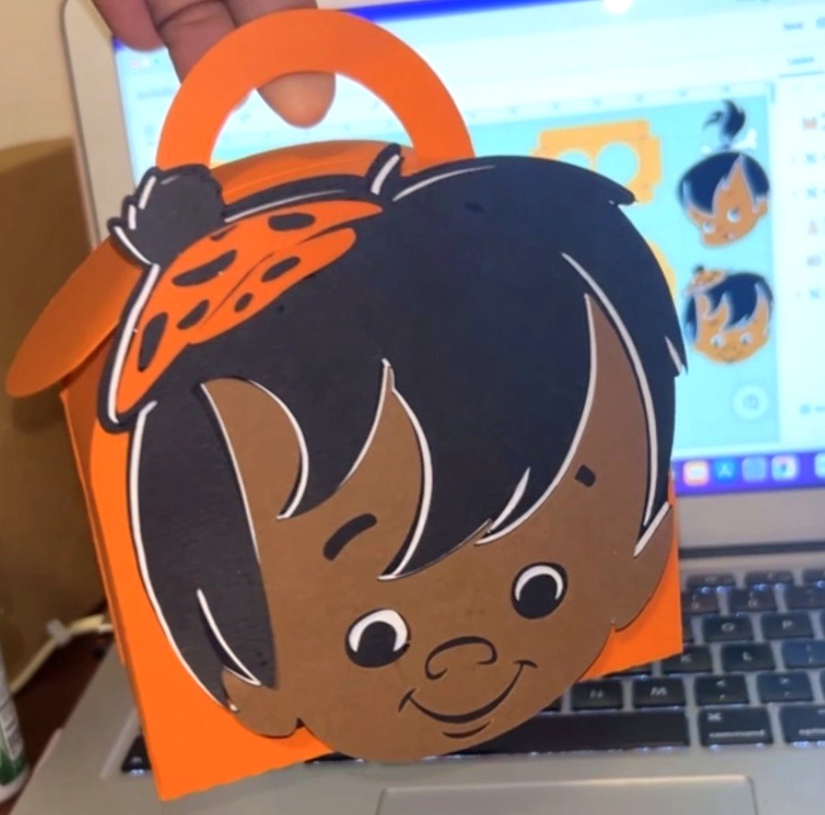 An orange favor box with a hand-cut layered paper face of a child, held up beside the cutting file on a laptop